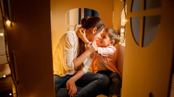 Young loving mother hugging with her little son in tent or carboard toy house at night. Concept of child loving parents and family having time together at night. — Stock Photo, Image