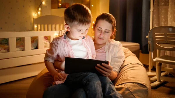Portrait of little boy sitting on mothers lap and watching cartoons on tablet computer at night. Concept of child education and family having time together at night