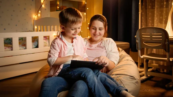 Uśmiechnięta matka z małym synkiem siedzącym w worku w nocy i śpiewającym tablet. Pojęcie wychowania dzieci i rodziny spędzających razem czas w nocy — Zdjęcie stockowe