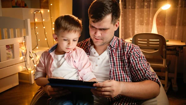 Muchacho sonriente con padre usando tableta en el dormitorio decorado para Navidad. Concepto de educación infantil y familia pasar tiempo juntos por la noche. — Foto de Stock