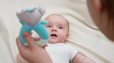 Genç anne küçük bebeğiyle yatakta konuşuyor ve renkli çıngırak oyuncaklarını sallıyor. Bebek eğitimi ve gelişimi kavramı.