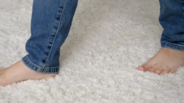 Pequeño chico descalzo en jeans caminando sobre una suave alfombra blanca en casa — Vídeo de stock