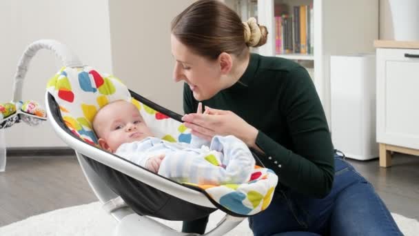 Portret van een lachende moeder naast een kleine baby die schommelt in een elektrische stoel. Ontwikkeling van het kind en gelukkig ouderschap — Stockvideo