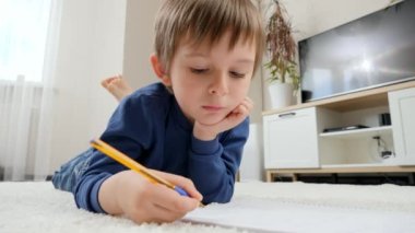 Küçük çocuk oturma odasındaki halıda yatarken ödevini defterden yapıyor. Yurtiçi eğitim ve çocuk gelişimi kavramı.