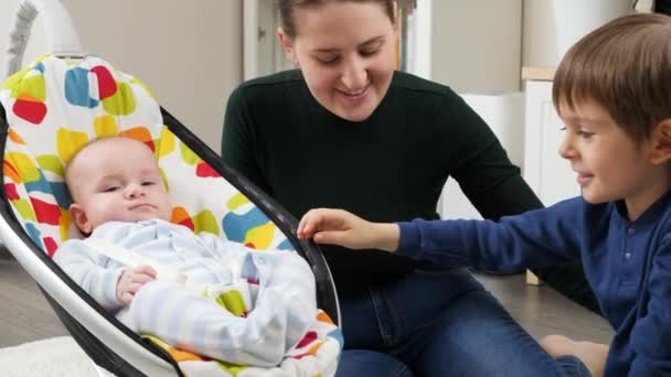 Hermano mayor y madre sonriente jugando con el pequeño niño meciéndose en el mecedero eléctrico. Desarrollo infantil y infancia feliz — Vídeos de Stock