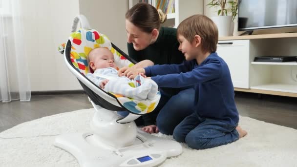 Mignon bébé garçon souriant basculant dans la chaise et regardant mère ond frère aîné. Développement de l'enfant et enfance heureuse — Video