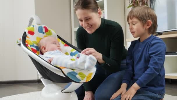 Feliz madre sonriente sentada en el suelo junto a su hijo mayor y su pequeño bebé meciéndose en una silla eléctrica. Desarrollo infantil y infancia feliz — Vídeos de Stock