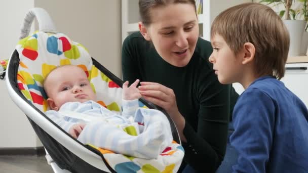 Starší bratr a usměvavá matka si povídají a hrají si s malým synem houpajícím se na dětském sedátku. Vývoj dítěte a šťastné dětství — Stock video