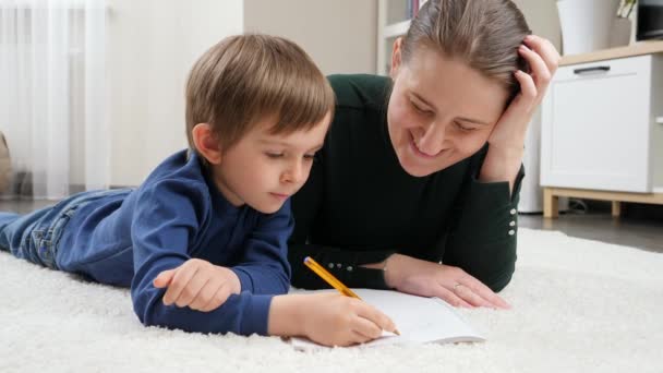Усміхнений хлопчик лежить на підлозі і робить домашнє завдання з мамою. Концепція виховання дітей, розвитку та щасливого виховання . — стокове відео