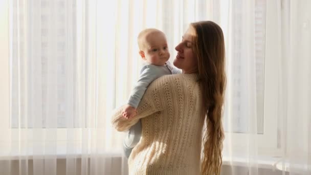 カーテン付きの大きな窓に立ってかわいい赤ちゃんと幸せな笑顔の若い母親。家族の幸せと子育ての概念 — ストック動画