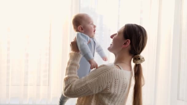 Joyeux rire tenant et jouant avec son bébé fils souriant contre le coucher du soleil dans une grande fenêtre. Concept de bonheur familial et de parentalité — Video