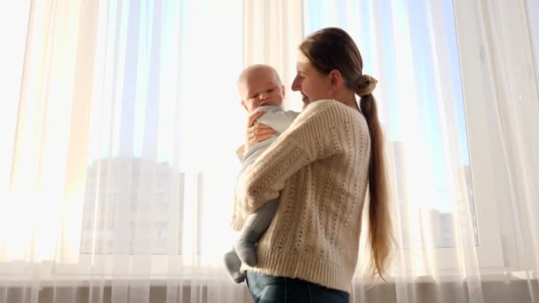 Retrato de una hermosa madre sosteniendo y besando a su pequeño hijo en una ventana grande. Concepto de felicidad familiar y paternidad — Vídeos de Stock