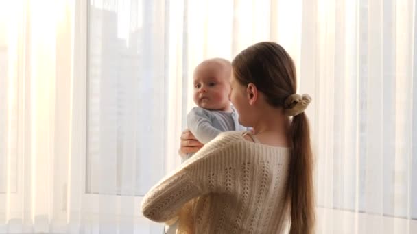 Lindo niño pequeño abrazando a su madre sonriente contra la gran ventana y la luz del atardecer. Concepto de felicidad familiar y paternidad — Vídeos de Stock