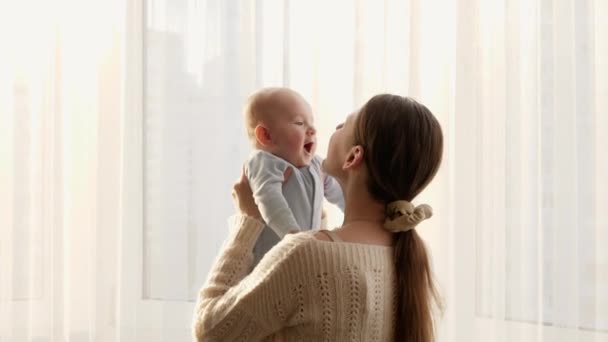 Щаслива усміхнена мати піднімається, грає і цілує свою дитину проти великого вікна вдома. Концепція сімейного щастя та виховання — стокове відео