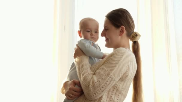 Retrato de una madre joven y cariñosa sonriendo a su pequeño hijo contra una gran ventana al atardecer. Concepto de felicidad familiar y paternidad — Vídeo de stock