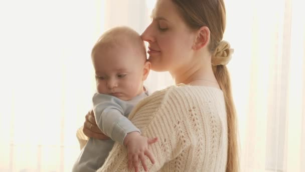 Ritratto di giovane madre sorridente con il piccolo bambino in piedi nella luce del tramonto che splende attraverso una grande finestra. Concetto di felicità familiare e genitorialità — Video Stock