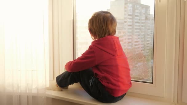 Küçük çocuk pencerenin kenarında oturuyor ve caddeye bakıyor ve günbatımı gökyüzünü pencereden izliyor.. — Stok video