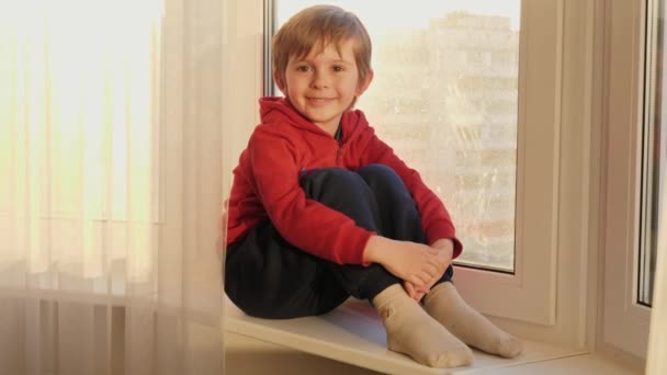 Portret uśmiechniętego chłopca siedzącego na parapecie i patrzącego w kamerę. Koncepcja pozostania w domu i pandemii covid-19 — Wideo stockowe