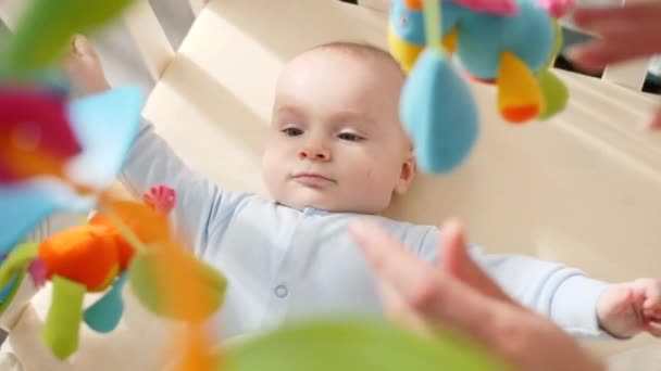 Kleines Baby, indem es mit buntem Spielzeug in der Krippe liegt und Mütter an der Hand hält. Konzept der Babyerziehung, Gesundheitsfürsorge und Entwicklung — Stockvideo