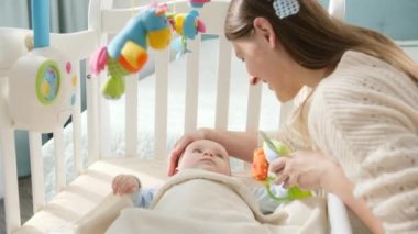 Küçük bebek beşikte yatıp peluş oyuncaklar ve genç bir anneyle oynuyor. Ebeveynlik kavramı, aile mutluluğu ve bebek gelişimi