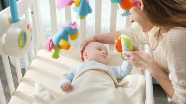 Joven madre jugando juguetes y acariciando a su pequeño hijo en la cuna. Concepto de paternidad, felicidad familiar y desarrollo del bebé — Vídeos de Stock