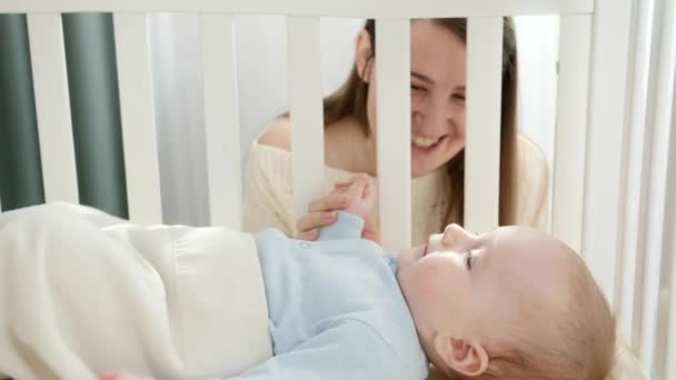 Feliz madre sonriente con un niño pequeño cogido de la mano y sonriéndose en una cuna de madera. Concepto de paternidad, felicidad familiar y desarrollo del bebé — Vídeos de Stock