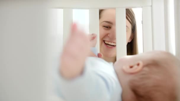 Portrait of happy laughing woman looking and holding her baby lying in crib. Concept of parenting, family happiness and baby development — Stock Video