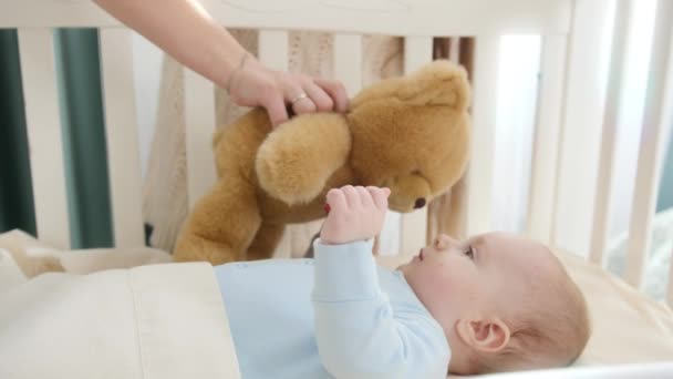 Imádnivaló kisfiú nyúlkál és játszik játékmackóval a bölcsőben. A szülőség, a család boldogsága és a baba fejlődése — Stock videók