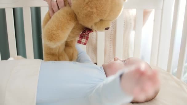 Jonge moeder speelt met teddybeer en haar kleine jongetje liggend in de wieg. Begrip ouderschap, gezinsgeluk en ontwikkeling van baby 's — Stockvideo