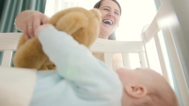 Щаслива усміхнена мати дарує плюшевому ведмедя своєму маленькому хлопчикові, який лежить у ліжку. Концепція виховання, сімейного щастя та розвитку дитини — стокове відео