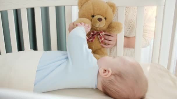 Іграшковий плюшевий ведмідь грає з маленьким хлопчиком, який лежить у колисці. Концепція виховання, сімейного щастя та розвитку дитини — стокове відео