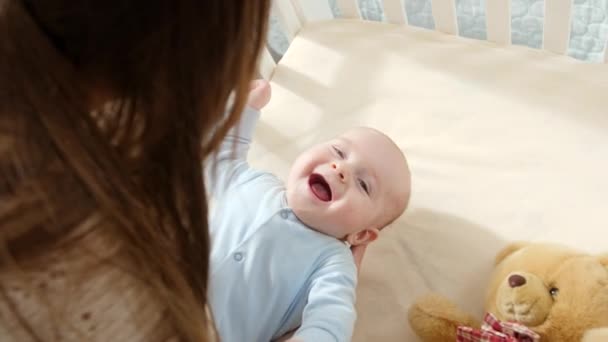 Fiatal anya fogja és ringatja mosolygós, aranyos kisfiát a bölcsőben. A szülőség, a család boldogsága és a baba fejlődése — Stock videók