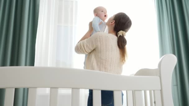 幼い母親はゆりかごの中に立ち、笑顔の赤ちゃんを抱いていた。子育て、家族の幸福と赤ちゃんの発達の概念 — ストック動画