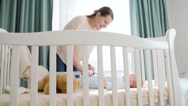 Sorridente giovane madre con il suo piccolo bambino sdraiato nella culla al mattino. Concetto di genitorialità, felicità familiare e sviluppo del bambino. — Video Stock