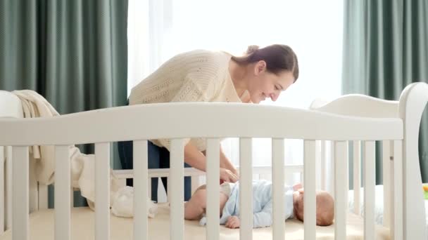 Schöne lächelnde Mutter, die ihren kleinen Sohn morgens in der Wiege auszieht, um Windeln zu wechseln. Konzept von Elternschaft, Familienglück und Babyentwicklung. — Stockvideo