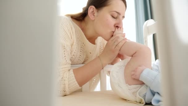Gelukkige lachende moeder die voeten kust van haar kleine baby liggend in de wieg. Begrip ouderschap, gezinsgeluk en ontwikkeling van baby 's. — Stockvideo