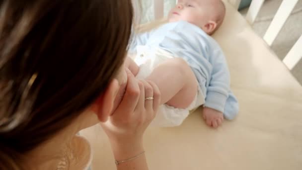 Mujer feliz mirando a su bebé tumbado en la cuna y besando pequeños pies. Concepto de paternidad, felicidad familiar y desarrollo del bebé. — Vídeo de stock