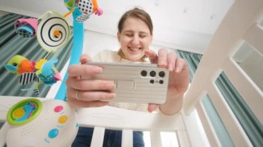 Mutlu gülümseyen anne bebeğinin sosyal medya için beşikte yattığını videoya çekiyor. Ebeveynlik kavramı ve çocuk tasviri