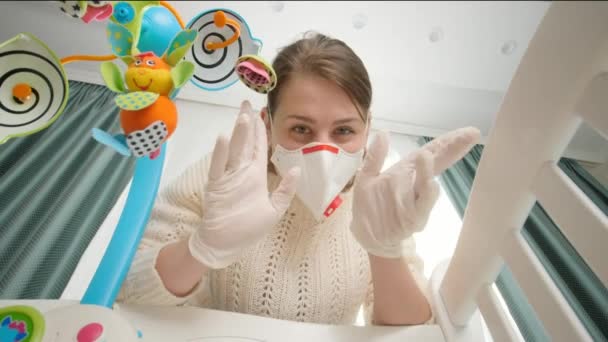Glückliche Frau mit medizinischer Maske, die mit Baby spielt, das in der Wiege liegt. Konzept der Kindererziehung und Babybetreuung während der Coronavirus-Covid-19-Pandemie. — Stockvideo