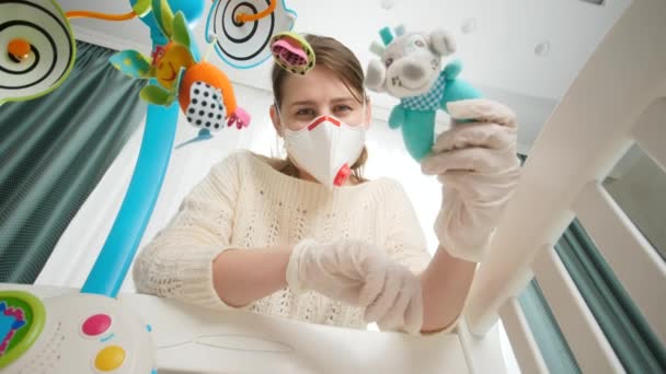 Giovane madre indossando maschera medica e guanti scuotendo giocattolo sonaglio con il suo piccolo bambino sdraiato nella culla. Concetto di genitorialità e cura del bambino durante la pandemia di coronavirus covid-19. — Video Stock