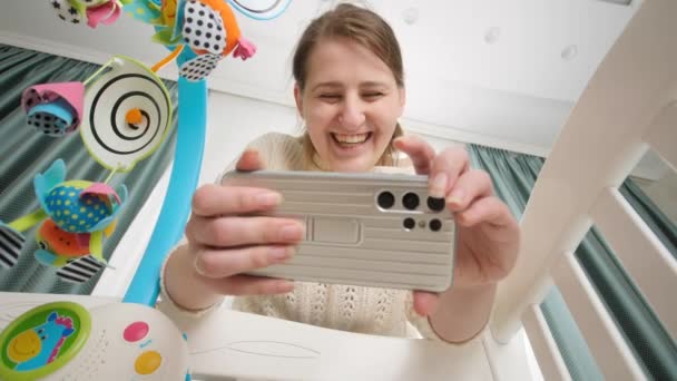 Glad smilende mor gør video på smartphone af hendes baby liggende i vugge. Begrebet forældre og gøre billeder af børn – Stock-video