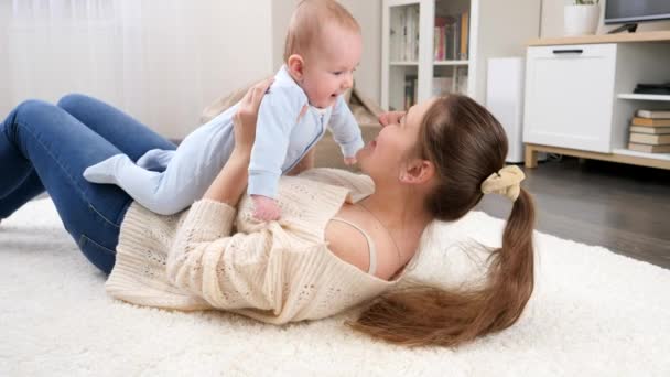 Gelukkige lachende moeder met kleine baby zoon liggend op de vloer in de woonkamer en plezier hebben. Begrip gezinsgeluk en ontwikkeling van kinderen — Stockvideo