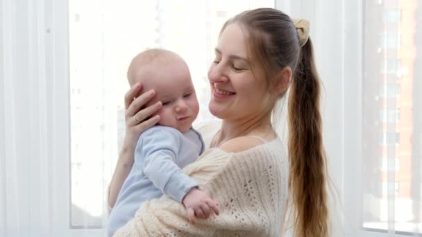 Retrato de una hermosa mujer sonriente abrazando y acariciando a su pequeño hijo en la ventana grande. Concepto de felicidad familiar y desarrollo infantil — Vídeo de stock