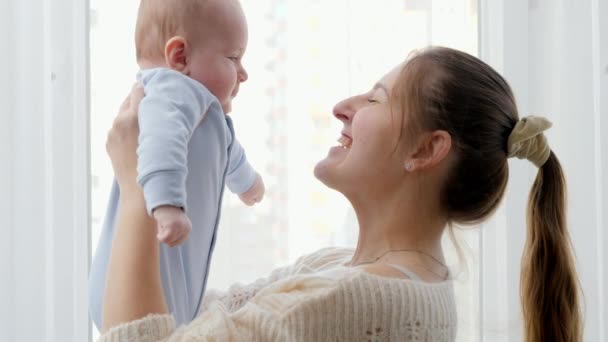Портрет молодої матері, яка піднімається і посміхається до свого маленького сина перед великим вікном вдома. Концепція сімейного щастя та розвитку дитини — стокове відео