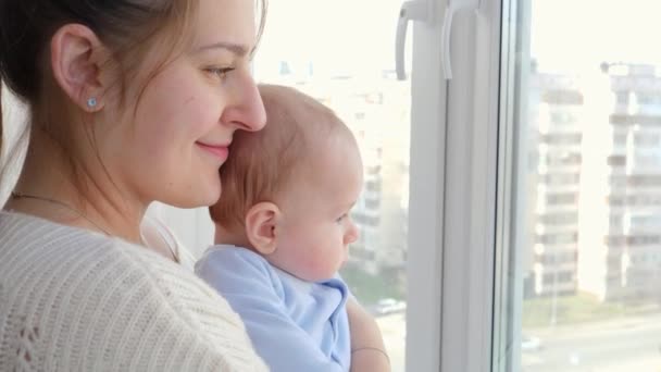 Retrato de una madre sonriente con un hijo pequeño mirando por la calle de la ciudad a través de la ventana. Concepto de felicidad familiar y desarrollo infantil — Vídeos de Stock