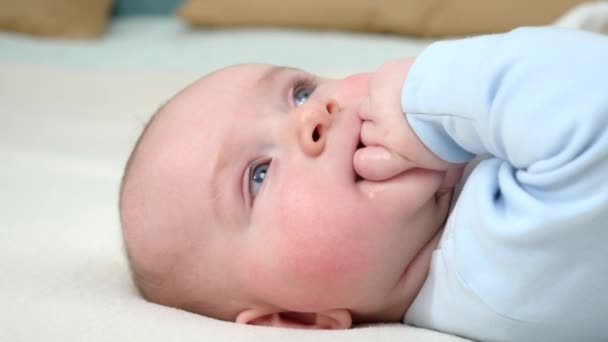 Retrato de niño adorable con ojos azules chupando su mano mientras está acostado en la cama. — Vídeo de stock