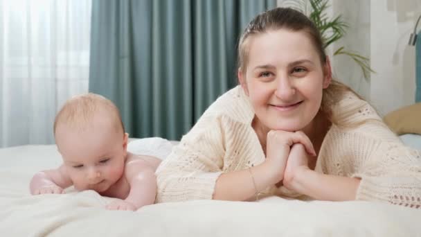 Młoda uśmiechnięta szczęśliwa matka leżąca z dzieckiem w łóżku i patrząca w kamerę. Pojęcie rodzicielstwa, opieki nad dziećmi i szczęścia rodzinnego — Wideo stockowe