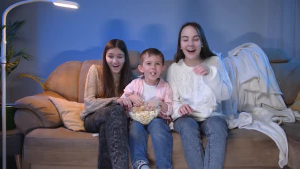 Leende liten pojke med två tonårstjejer tittar på TV sent på kvällen och äter popcorn från stora skålen — Stockvideo