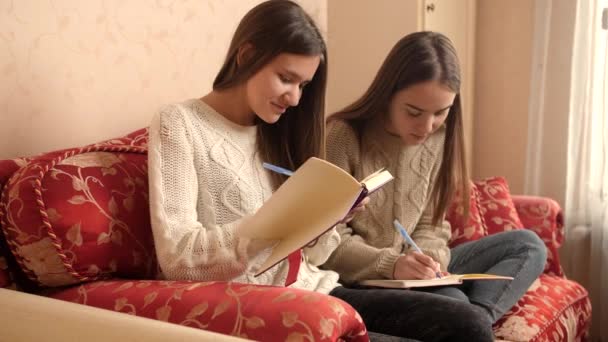 Két boldog mosolygós lány mosolyog és nevet, miközben házit csinálnak és naplót vagy naplót írnak. — Stock videók