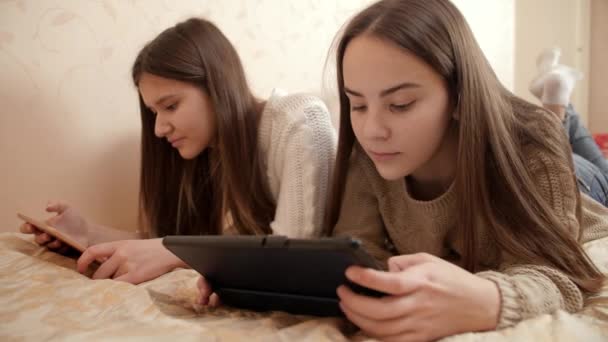 Портрет двух улыбающихся девочек-подростков, лежащих на кровати и использующих планшетные компьютеры. Современное образование и подростки с использованием гаджетов — стоковое видео